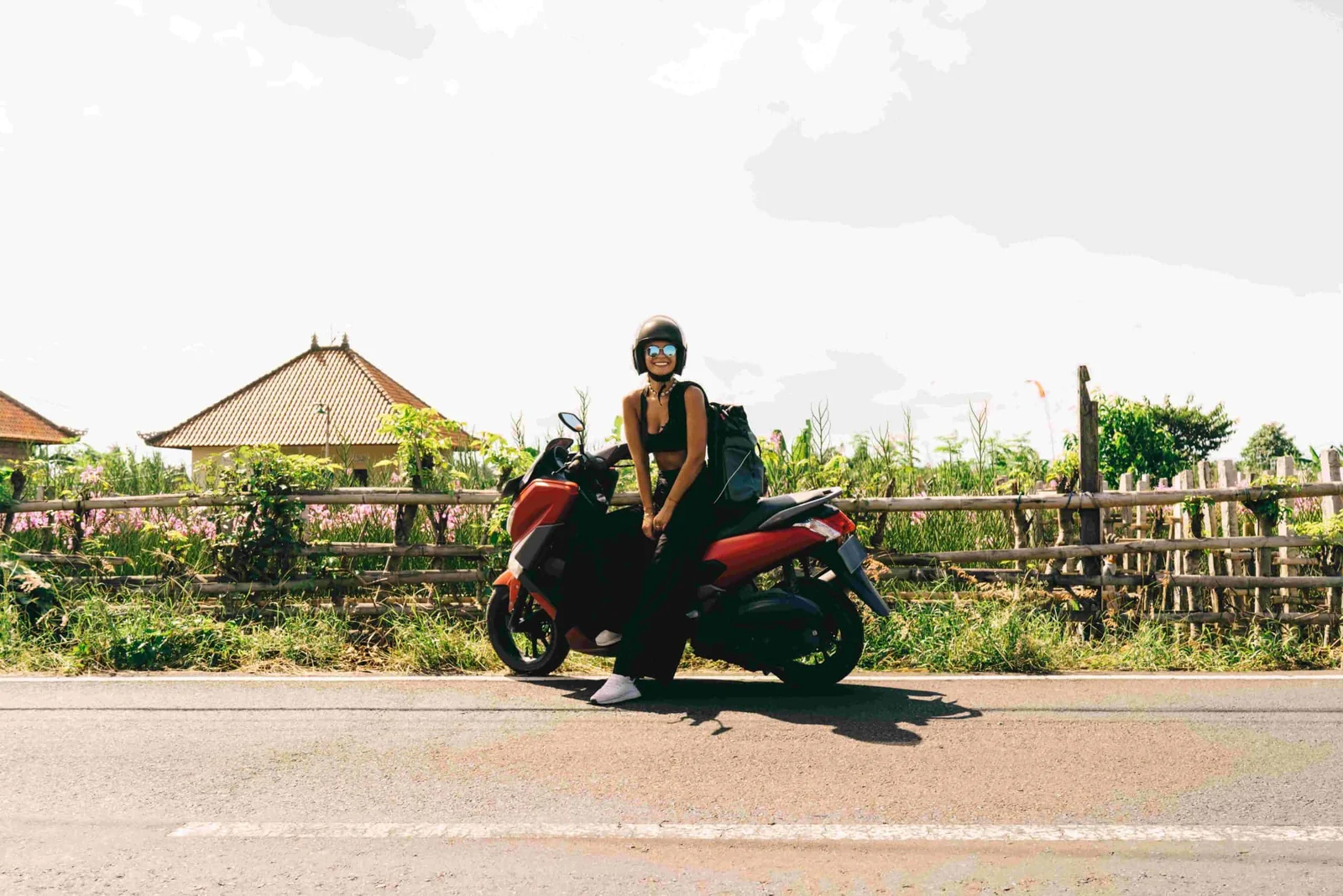 Seorang traveler perempuan menikmati liburan di destinasi eksotis Bali dengan duduk di atas motor sewaan di pedesaan Ubud, Bali. Latar belakang rumah tradisional dan alam hijau menambah suasana tropis, cocok untuk eksplorasi dengan sewa motor Bali.