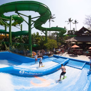 Beberapa pengunjung bermain selancar di wahana FlowRider Waterbom Bali, dengan latar belakang seluncuran air hijau yang ikonik dan suasana taman tropis yang ramai.