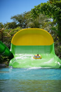 Seorang anak sedang meluncur di wahana Mini Boomerang Waterbom Bali menggunakan ban pelampung kuning, dikelilingi pepohonan tropis dan langit biru cerah.