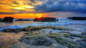 Pemandangan matahari terbenam yang memukau di batu bolong beach, Bali, dengan awan dramatis, ombak yang menghantam, dan bebatuan berlumut di tepi pantai.