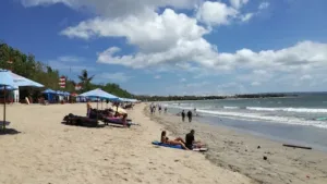 Suasana siang hari di Kuta Beach Bali dengan wisatawan berjemur di bawah payung pantai, menikmati pasir putih dan pemandangan laut biru yang tenang.