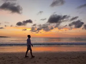 Seorang pria berjalan di tepi Kuta Beach Bali saat matahari terbenam, dengan langit berwarna jingga dan suasana pantai yang tenang.