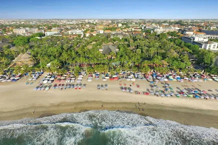 Pemandangan udara Pantai Legian di Bali dengan deretan payung pantai warna-warni, pasir keemasan, ombak laut, dan latar belakang pepohonan hijau serta bangunan kota.