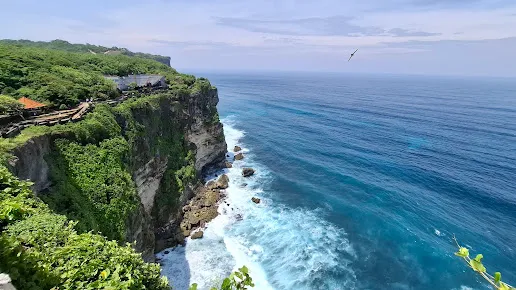 Tebing tinggi Uluwatu Temple Bali dengan pemandangan laut biru jernih dan pepohonan hijau yang rimbun, menawarkan panorama alam yang spektakuler di ujung selatan pulau.